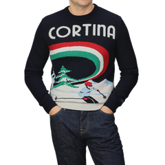 A person is wearing the Navy Blue Wool Cashmere Cortina Sweater from MC2 Saint Barth, featuring a skier and "Cortina" text design, with their hands in pockets while also wearing jeans.