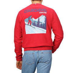 A person is sporting the Bright Red Cotton Courchevel Sweater by MC2 Saint Barth, featuring a skiing graphic and "COURCHEVEL ADDICTED" text on the back, embodying the perfect après-ski vibe, paired with blue jeans.