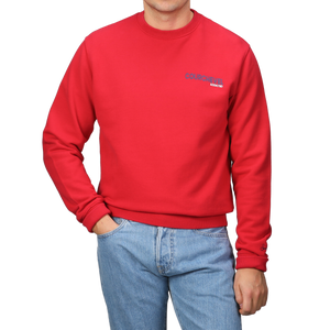 Wearing an MC2 Saint Barth Bright Red Cotton Courchevel Sweater adorned with text and blue jeans, a person stands against a gray background, perfectly capturing the relaxed spirit of Courchevel's après-ski scene.