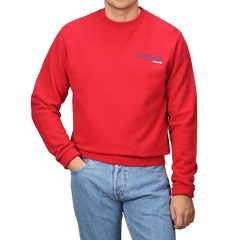 Wearing an MC2 Saint Barth Bright Red Cotton Courchevel Sweater adorned with text and blue jeans, a person stands against a gray background, perfectly capturing the relaxed spirit of Courchevel's après-ski scene.