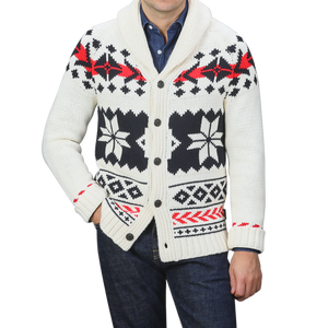 A person is wearing an MC2 Saint Barth Cream White Wool Nordic Aztec Cardigan with a shawl collar, showcasing a black and red geometric pattern against a cream base, elegantly layered over a blue shirt and dark jeans.