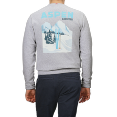 Someone is wearing the Grey Melange Cotton Aspen Sweater by MC2 Saint Barth, featuring a graphic of snowy mountains, trees, and ski poles. The phrase "Aspen Addicted" above the image perfectly captures the essence of Aspen ski resorts, ideal for those "Aprés Ski" moments.