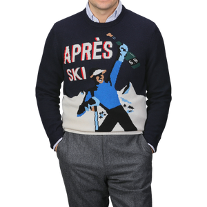 A person wearing the MC2 Saint Barth Navy Blue Wool Cashmere Aprés Ski Sweater, adorned with a graphic of a skier holding a bottle and majestic ski resorts in the background.