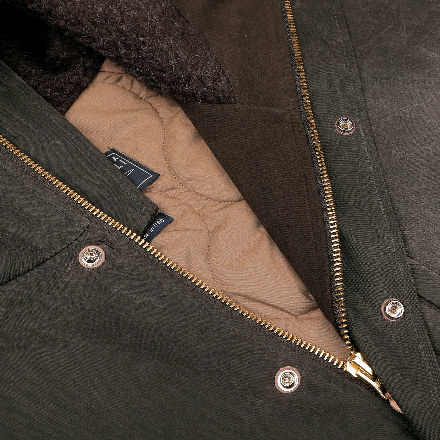 Close-up of the Dark Green Waxed Cotton Mountain Parka by Manifattura Ceccarelli, featuring a gold zipper and brown quilted lining. The jacket showcases metal snap buttons and a woolen collar, with a partially visible fabric tag displaying the logo inside.