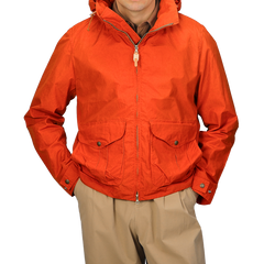 A person is wearing the Manifattura Ceccarelli Orange Cotton Nylon Ripstop All Seasons Blazer Coat with a hood, zipper, and two front pockets over tan pants. The jacket boasts a detachable lining for versatility in different weather conditions.