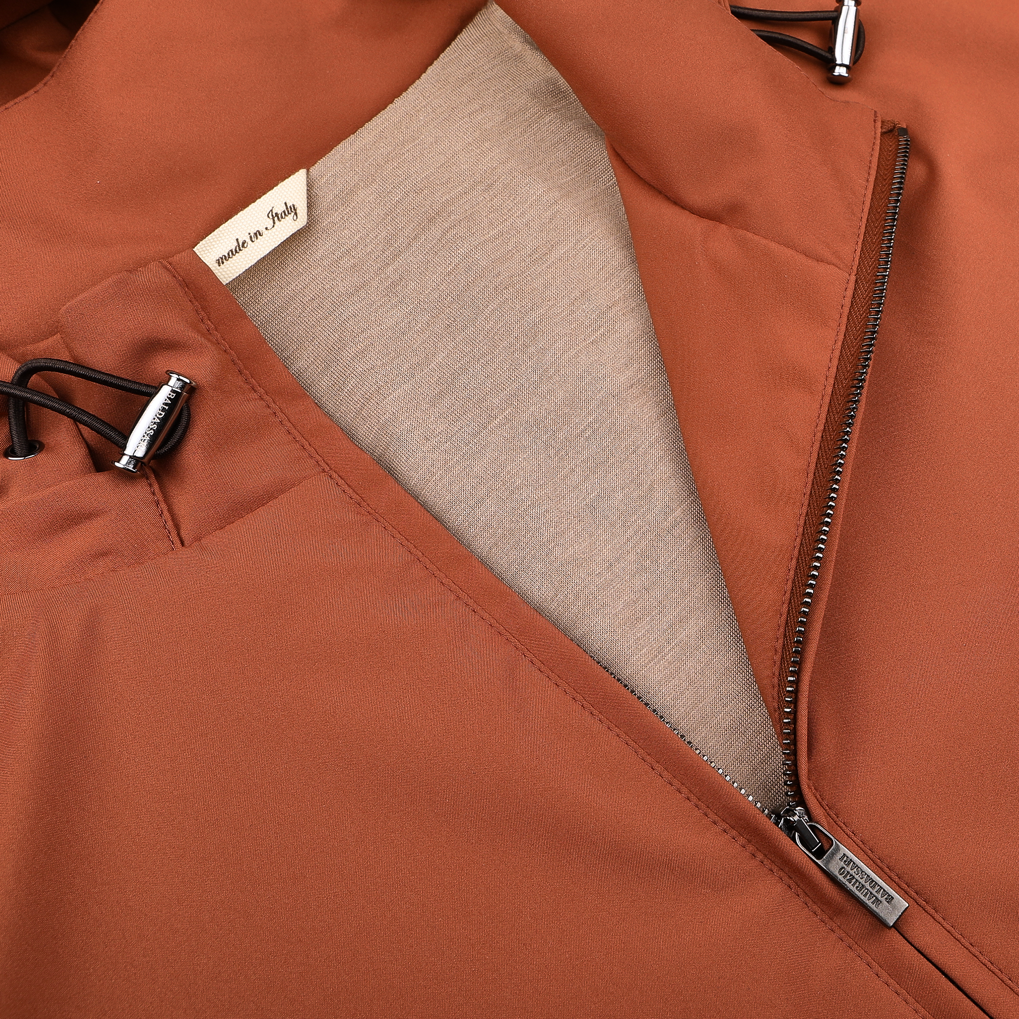 Close-up of the Terracotta Brown Nylon Hooded Blouson Jacket by Maurizio Baldassari, showcasing a partially opened zipper revealing a beige lining and adjustable drawstrings with metal toggles. The "Made in Italy" tag is visible on this technical nylon piece.