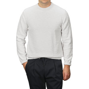 A person wearing a Maurizio Baldassari Cream Summer Mouline Crew Neck Sweater and black pants stands against a gray background, with hands partially in their pockets, highlighting the comfortable cotton-viscose blend.