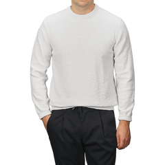 A person wearing a Maurizio Baldassari Cream Summer Mouline Crew Neck Sweater and black pants stands against a gray background, with hands partially in their pockets, highlighting the comfortable cotton-viscose blend.