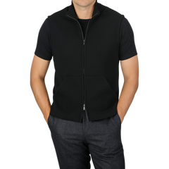 Here’s a revised version of the sentence incorporating the given product data:

A person wearing a black short-sleeve shirt under the Maurizio Baldassari Black Milano Stitch Wool Zip Gilet pairs it with dark gray pants, with hands partially in the pants pockets. The background is plain light gray.