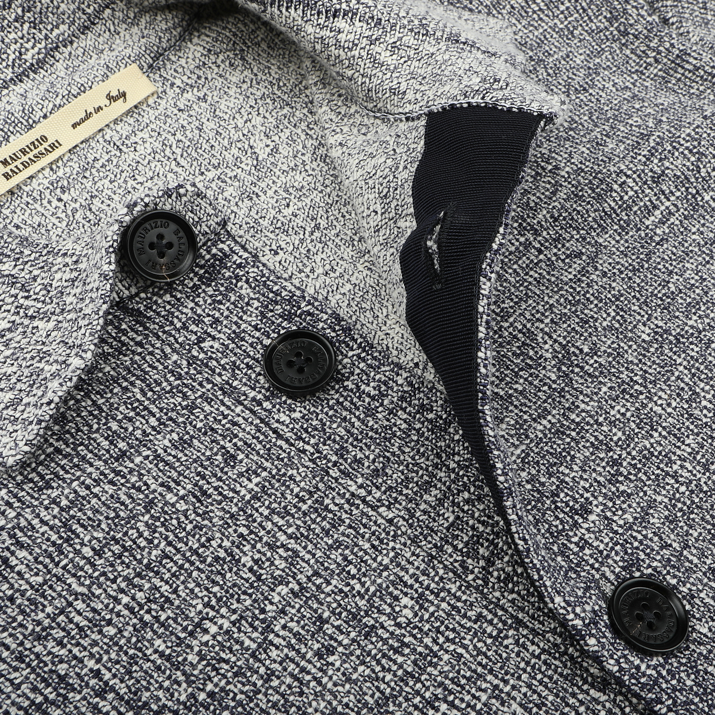 Close-up of a textured gray jacket featuring three black buttons and a visible "Made in Italy" label. Crafted by Maurizio Baldassari, this piece echoes the elegance and craftsmanship of the Blue Melange Summer Mouline Blouson.