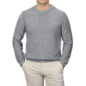 A person wearing a Blue Melange Summer Mouline Crew Neck Sweater by Maurizio Baldassari and beige pants stands with hands in pockets against a plain background.