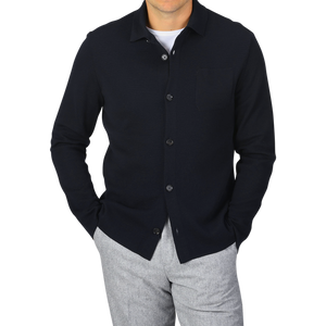 A person wearing a Navy Blue Wool Milano Knitted Overshirt by Maurizio Baldassari with a pocket on the left side and light grey pants, with hands in pockets, standing against a plain grey background.