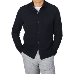 A person wearing a Navy Blue Wool Milano Knitted Overshirt by Maurizio Baldassari with a pocket on the left side and light grey pants, with hands in pockets, standing against a plain grey background.