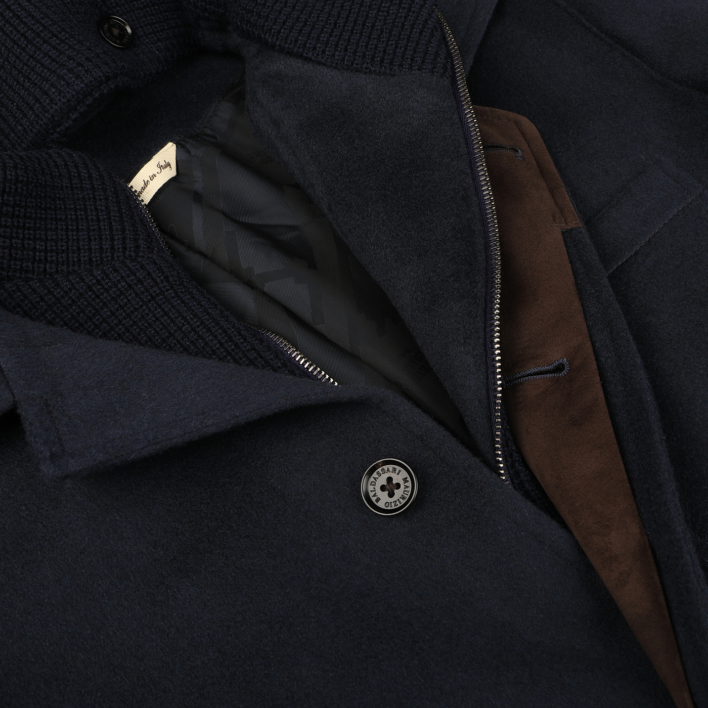 Close-up of the Maurizio Baldassari Navy Cashmere Half Padded Chore Coat, showcasing a partially visible brown suede trim and interior lining. A metal button embossed with the brand's logo stands out, while the Thermore technical down adds warmth to this sophisticated garment.