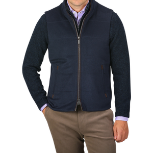 A person wearing a Navy Water Repellent Pure Cashmere Gilet by Maurizio Baldassari over a blue sweater and light purple shirt, with brown pants. The person has their hands in their pockets against a plain background.