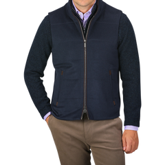 A person wearing a Navy Water Repellent Pure Cashmere Gilet by Maurizio Baldassari over a blue sweater and light purple shirt, with brown pants. The person has their hands in their pockets against a plain background.