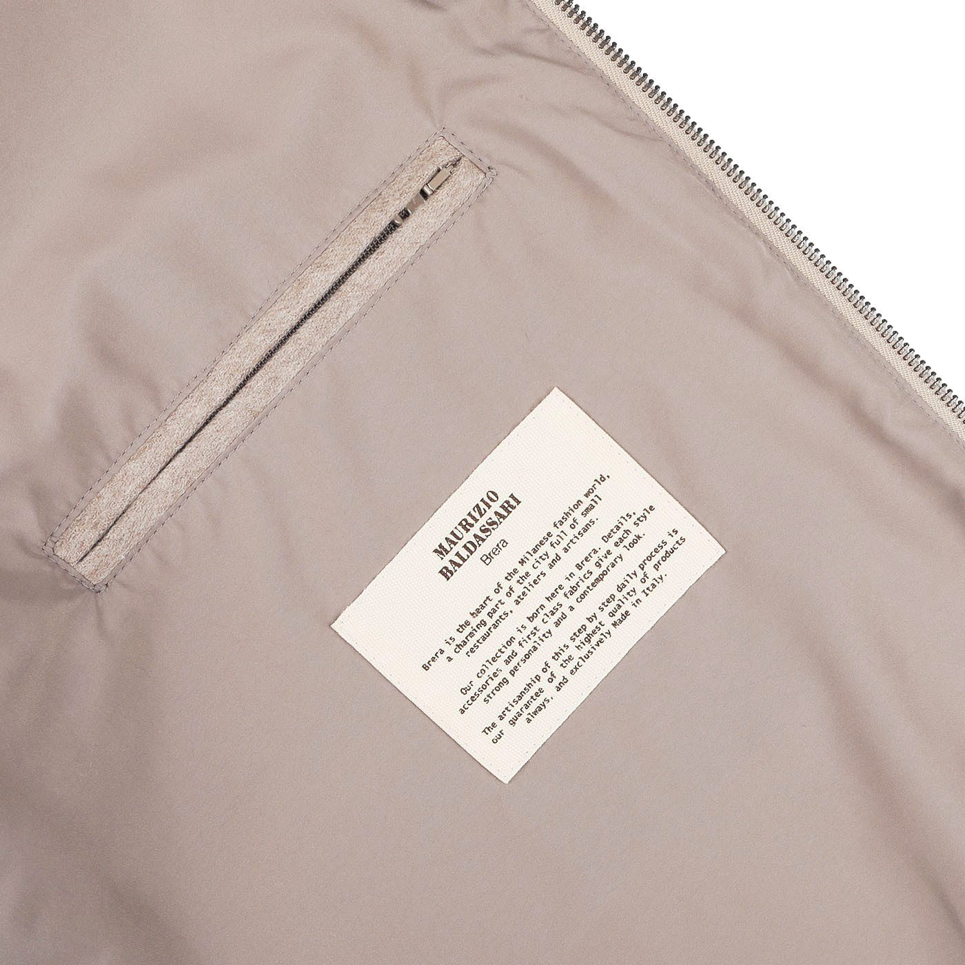 The Oatmeal Beige Cashmere Zip Blouson by Maurizio Baldassari features a beige fabric with a small zippered pocket and a sewn-in label printed in black text that provides manufacturing and care details, reminiscent of the intricate detailing on the jacket.