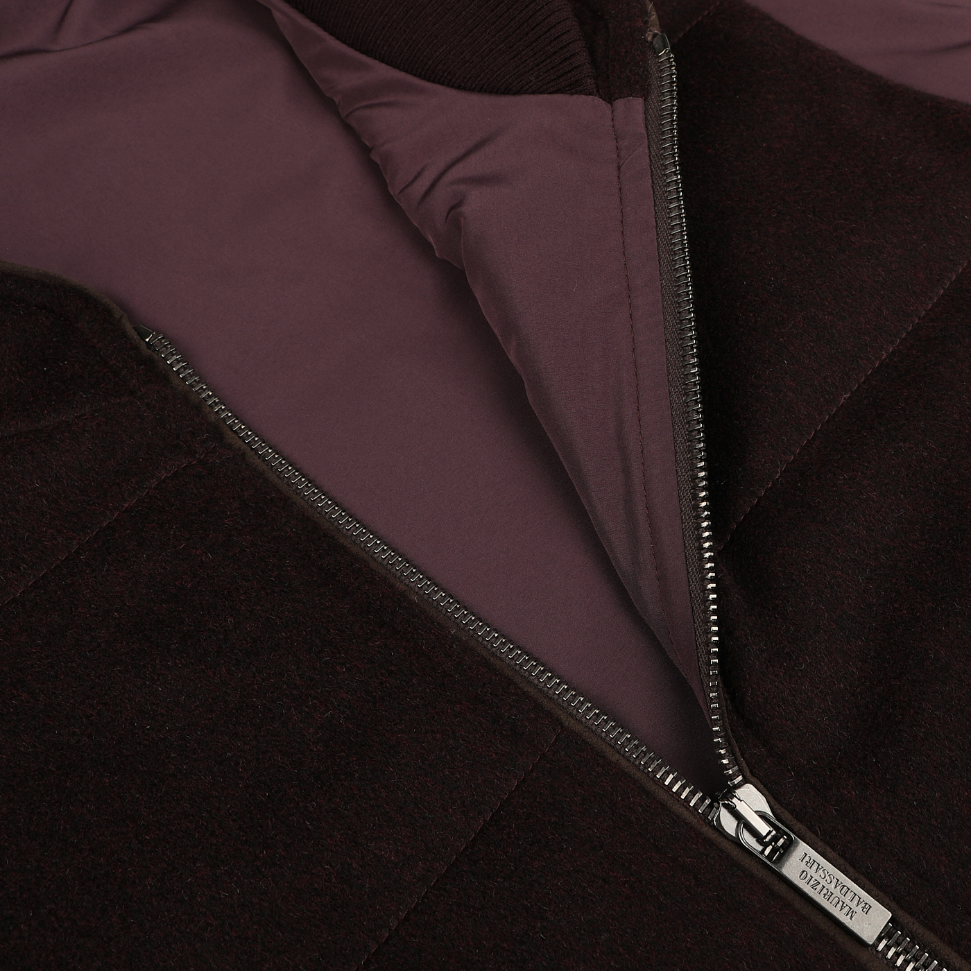 Close-up of the Plum Water Repellent Cashmere Gilet by Maurizio Baldassari, showcasing its maroon and black design with a water-repellent finish. The metal zipper is partially open, revealing the inner lining, and the zipper pull features engraved text.