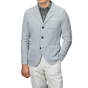 A man exudes relaxed luxury in the Sky Blue Summer Mouline Knitted Jacket by Maurizio Baldassari, paired with a dark gray collared shirt and white pants, one hand casually in his pocket. The cotton-viscose blend enhances sophistication against the plain background.