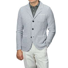 A man exudes relaxed luxury in the Sky Blue Summer Mouline Knitted Jacket by Maurizio Baldassari, paired with a dark gray collared shirt and white pants, one hand casually in his pocket. The cotton-viscose blend enhances sophistication against the plain background.