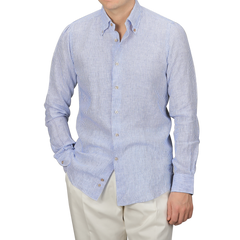 Wearing Mazzarelli's Blue Striped Organic Linen BD Shirt with the top button undone and paired with white pants, a person stands casually with one hand in their pocket.