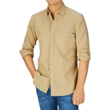 A man wearing a Khaki Beige Cotton Gabardine Regular Fit Shirt by Italian shirtmaker Mazzarelli and stone-washed blue jeans against a blue background.