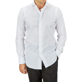 A man wearing a crisp Mazzarelli white cotton twill cutaway slim shirt and dark trousers, standing against a grey background. Only his torso is visible in the image.