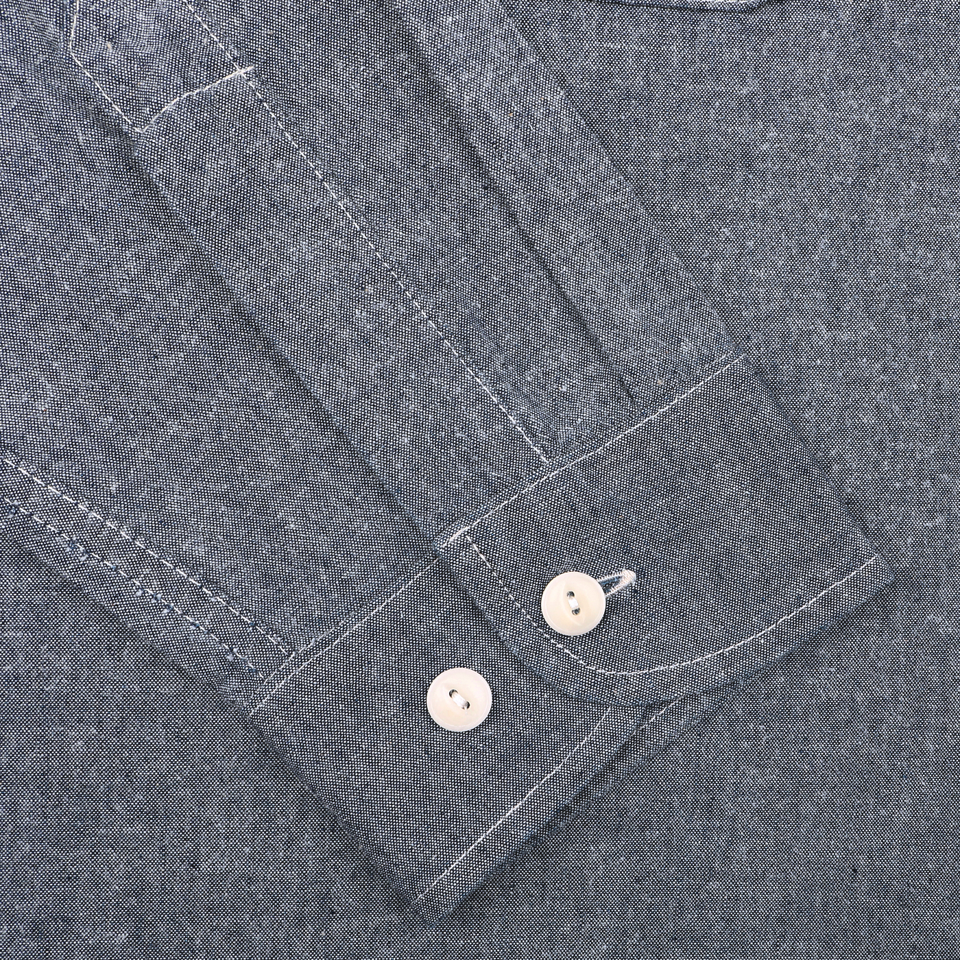 Close-up of the versatile Denim Blue Cotton Chambrey 06 CH Shirt by Merz b. Schwanen, featuring a gray sleeve with white buttons and visible stitching against a textured background.