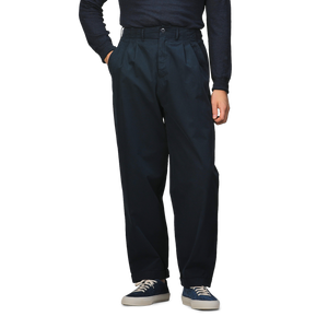 A person stands with hands in pockets wearing Merz b. Schwanen Navy Blue Cotton Poplin 03 Pants, a dark long-sleeve shirt crafted from organic cotton for added comfort, blue and white sneakers.