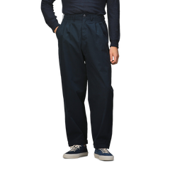 A person stands with hands in pockets wearing Merz b. Schwanen Navy Blue Cotton Poplin 03 Pants, a dark long-sleeve shirt crafted from organic cotton for added comfort, blue and white sneakers.