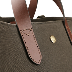 Close-up of the Army Cuoio M/S Canvas Verve Tote Bag by Mismo, showcasing a brown leather handbag strap with stitching and a round gold emblem, highlighting exquisite leather details against the brown fabric.