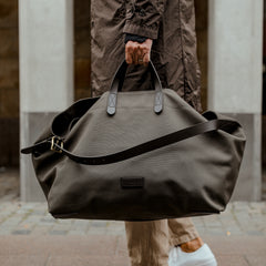 Person holding a large, Army Dark Brown M/S Haven Weekend Bag by Mismo by the handles while walking on a city street.