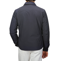 Against a plain backdrop, someone models Montecore's minimalist elegance by wearing the Dark Blue Light Down Waterproof Shirt Jacket with white pants, their back to the camera.