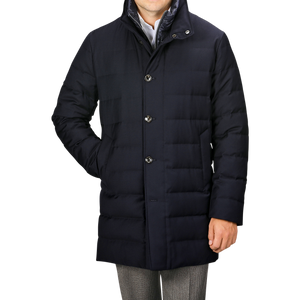 A person wearing a Navy Blue Super 120s Wool Batavia Padded Coat by Montecore, featuring a high collar and button-up design, paired with grey trousers, standing with one hand relaxed by their side.