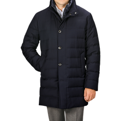 A person wearing a Navy Blue Super 120s Wool Batavia Padded Coat by Montecore, featuring a high collar and button-up design, paired with grey trousers, standing with one hand relaxed by their side.