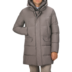 A man wearing the Taupe Beige Nylon Down Padded Parka by Montecore, which features knee-length padding, a front zipper, and two large front pockets, stands with one hand in a pocket.