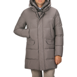 A man wearing the Taupe Beige Nylon Down Padded Parka by Montecore, which features knee-length padding, a front zipper, and two large front pockets, stands with one hand in a pocket.