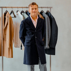 A person stands in front of a clothing rack filled with various coats. They are wearing a Navy Blue Loro Piana Wool Tailored Coat from Baltzar Sartorial, paired with a light shirt.