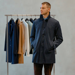 A man in a dark coat stands with hands in pockets before a clothing rack laden with stylish car coats and jackets, including the Navy Blue Nylon Thermore Padded Car Coat by Baltzar Sartorial, which is crafted from technical nylon and features Thermore Ecodown lining for added warmth.