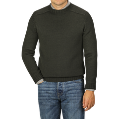 A man wearing a Nitto Khaki Green Merino Wool Garbis Sweater and blue jeans stands against a plain grey background, with only his torso and part of his arms visible.