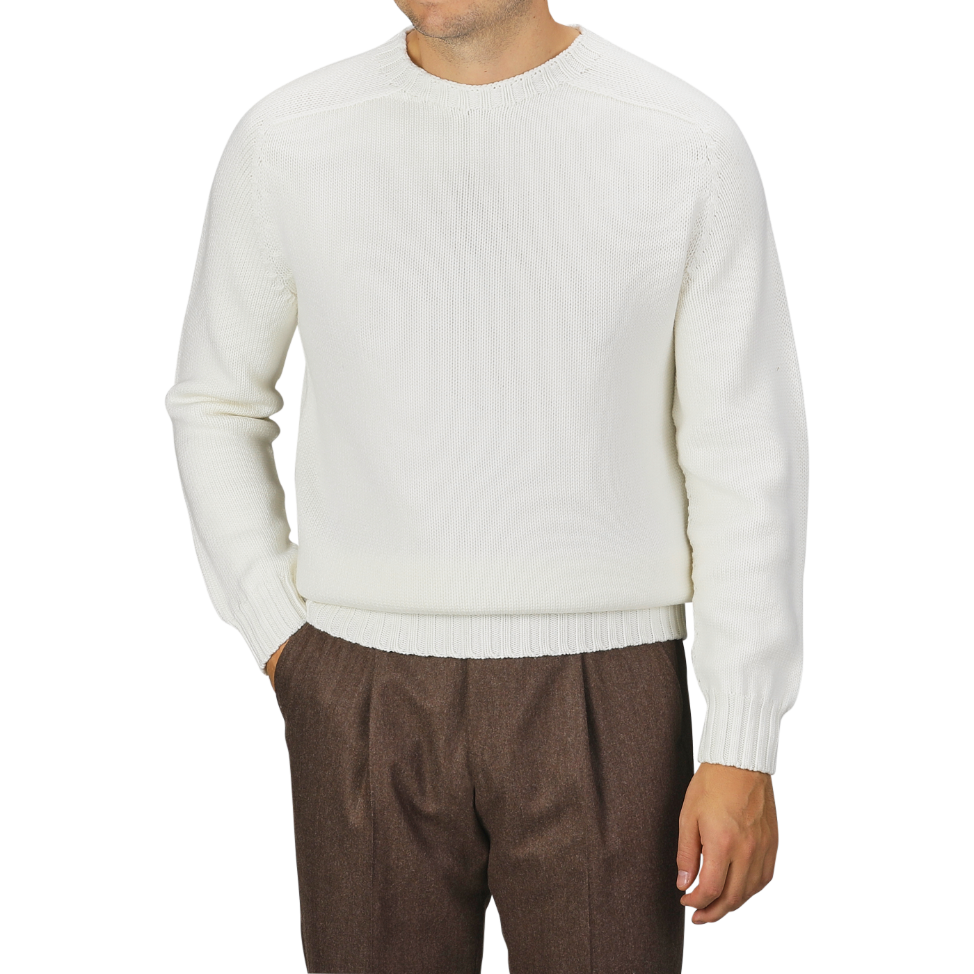 A man is wearing an Ecru White Merino Wool Garbis Sweater by Nitto and brown trousers. His arms are relaxed at his sides with hands in his pockets. The background is plain and gray.