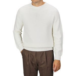 A man is wearing an Ecru White Merino Wool Garbis Sweater by Nitto and brown trousers. His arms are relaxed at his sides with hands in his pockets. The background is plain and gray.