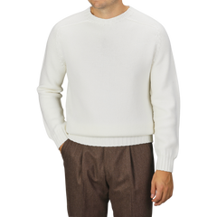 A man is wearing an Ecru White Merino Wool Garbis Sweater by Nitto and brown trousers. His arms are relaxed at his sides with hands in his pockets. The background is plain and gray.