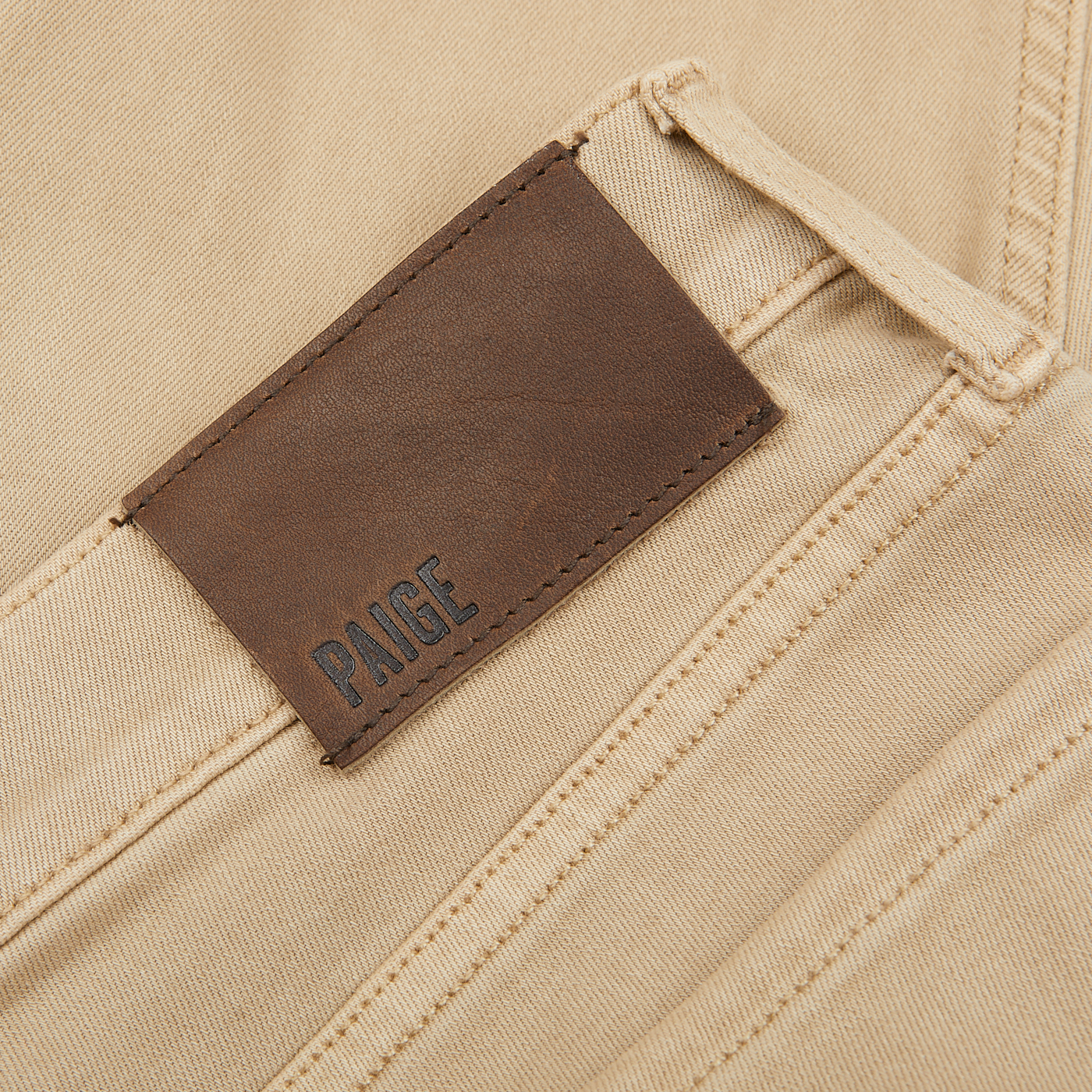 Close-up of a brown leather label with the word "Paige" embossed on a pair of Caramel Beige Cotton Transcend Federal Slim Jeans, made with Transcend fabric for a comfortable fit.