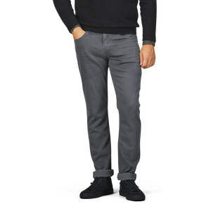 A person wearing a black long-sleeve shirt, black shoes, and Paige Charcoal Cotton Transcend Federal Slim Jeans in onyx grey stands with hands in front pockets. The jeans, made from comfortable Transcend fabric, are rolled up at the cuffs. The head and upper body are not visible.