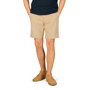 A person standing with their hands behind their back, wearing a navy blue t-shirt, Paige Khaki Beige Cotton Blend Phillips shorts, and light brown shoes.