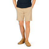 A person standing with their hands behind their back, wearing a navy blue t-shirt, Paige Khaki Beige Cotton Blend Phillips shorts, and light brown shoes.
