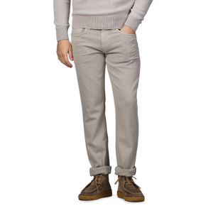 A person poses against a gray background, wearing a beige sweater, Paige Moroccan Clay Cotton Transcend Federal Slim Jeans, and brown shoes.