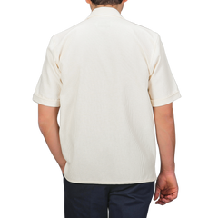 A person wearing a Peregrine Cream Dry Cotton Cuban Collar Shirt with dark pants stands back to the camera against a gray background, highlighting the pure cotton fabric.