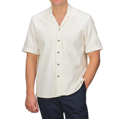 An individual stands against a gray backdrop, wearing Peregrine's Cream Dry Cotton Cuban Collar Shirt paired with dark pants.